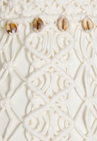 Bílá macramé látka s propracovanými uzlovými vzory. Vrchní část je ozdobena třemi dekorativními hnědými mušlemi navlečenými podélně.