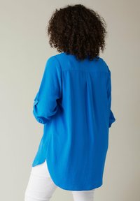 Person med lockigt hår som bär en ljusblå långärmad tröja och vita byxor, står med ryggen vänd mot betraktaren mot en plain bakgrund.