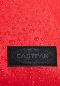 Červená voděodolná látka má na sobě kapky vody. V dolním rohu se nachází černý obdélníkový štítek s reliéfním nápisem "EASTPAK".