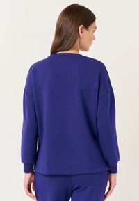 Felpa viola a maniche lunghe con una vestibilità rilassata, colletto e polsini a costine. Texture liscia, caratterizzata da un colore uniforme senza motivi.