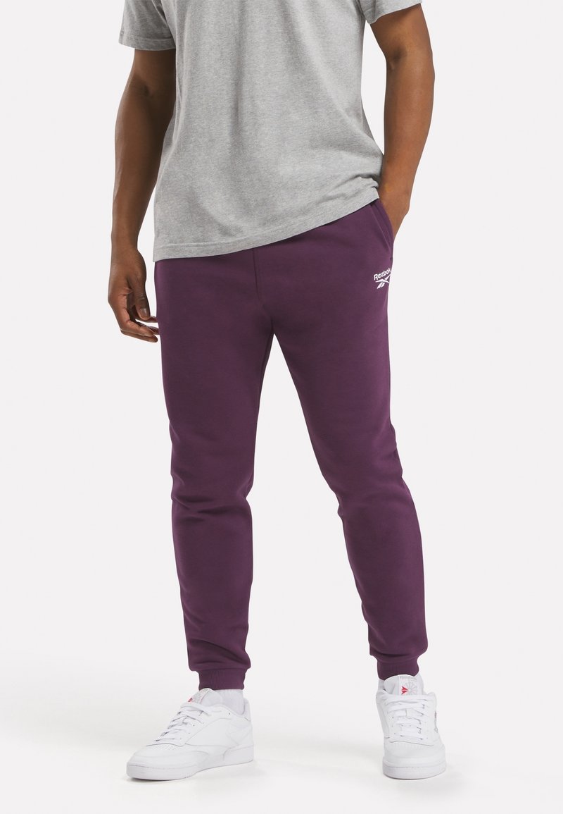 Purple sweatpants with cuffed ankles, a simple design, and a white logo on the left thigh, paired with a gray T-shirt and white sneakers.