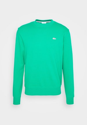 Green sweatshirt with a round neckline and a toggle drawstring; cream-colored trousers; white sneakers with light gray accents.