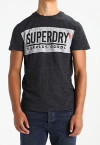 Šedé tričko s krátkým rukávem a bílým horizontálním pruhem, na kterém je text "SUPERDRY SURPLUS GOODS". Měkký bavlněný materiál, volný střih.