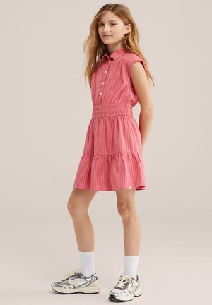 Fetiță tânără în picioare, purtând o rochie roz fără mâneci cu dungi albe, șosete albe și adidași argintii, pe un fundal simplu.