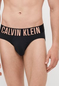 Culottes en coton noir avec une large ceinture élastique portant l'inscription "CALVIN KLEIN" en rose clair. Texture lisse et taille ajustée.