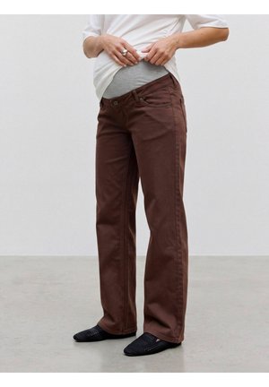 Personne portant un pantalon de maternité marron et des chaussures noires à enfiler, relevant une chemise blanche pour révéler un panneau de maternité gris.