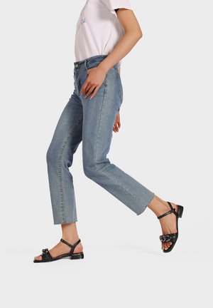 Sandali flat neri con cinturino a fibbia e accentuazione decorativa a fiocco, abbinati a jeans in denim azzurri a vita alta con orlo non rifinito.