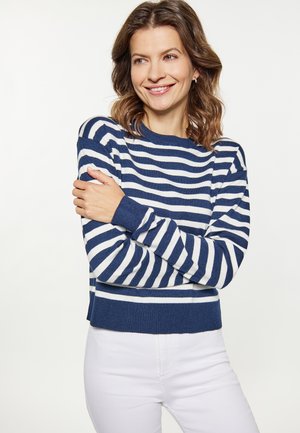 Femme souriante aux cheveux bruns ondulés portant un pull rayé bleu marine et blanc et un pantalon blanc, bras croisés sur fond blanc.