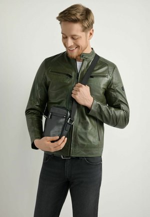 Uśmiechnięty mężczyzna w zielonej skórzanej kurtce trzyma czarną torbę crossbody z zamkami błyskawicznymi na jasnym tle.