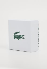 Baltasis tekstūruotas dėžutė su žaliu krokodilo logotipu priekyje ir užrašu "LACOSTE" atspausdintu vertikaliai šone.