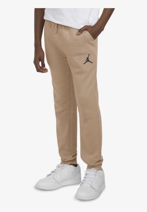 Osoba ubrana w beżowe spodnie dresowe z czarnym logo Jumpman na udzie, białe sneakersy i białą koszulkę, stoi z jedną ręką w kieszeni.