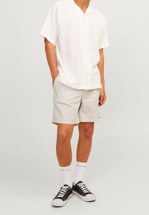 Homme portant une chemise blanche à manches courtes boutonnée, un short beige clair, des chaussettes blanches montant jusqu’au mollet et des baskets basses noires, debout, les mains détendues.