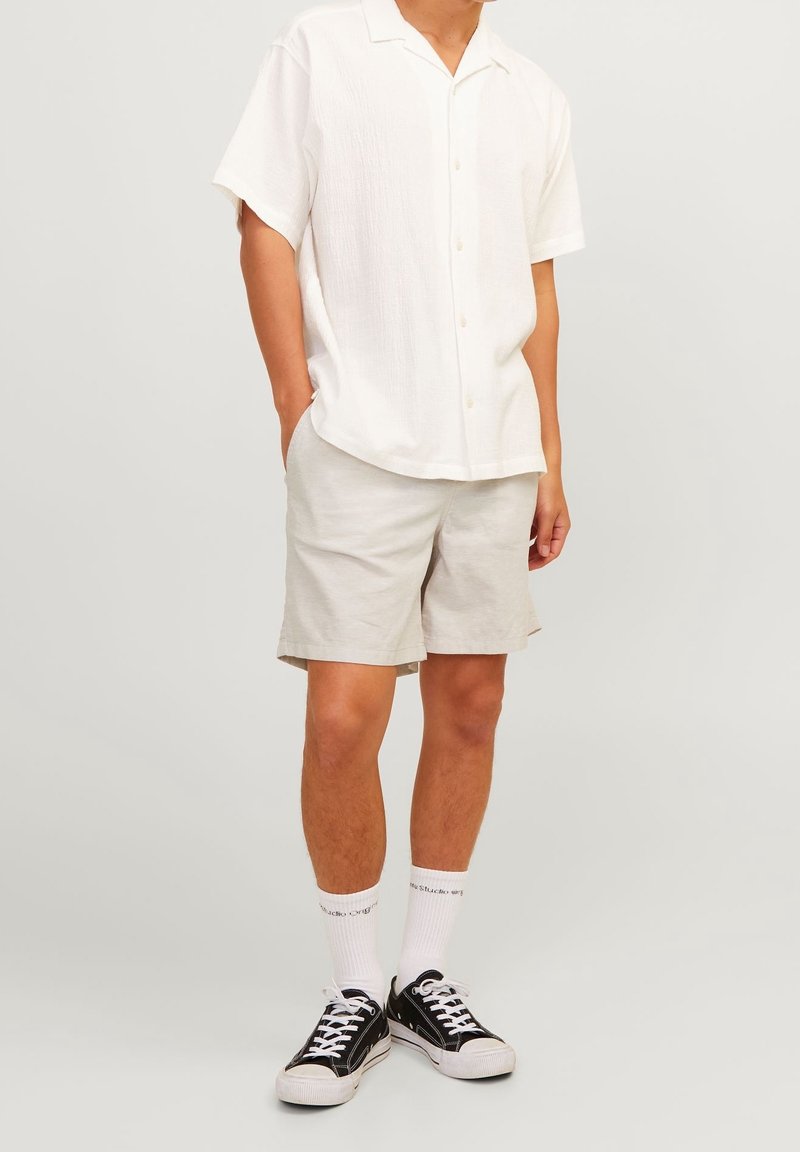 Homme portant une chemise blanche à manches courtes boutonnée, un short beige clair, des chaussettes blanches montant jusqu’au mollet et des baskets basses noires, debout, les mains détendues.