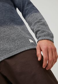 Homme portant un pull en maille gris dégradé à manches longues avec une étiquette blanche près de l'ourlet, associé à un pantalon marron foncé, main détendue le long du corps.
