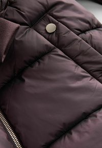 Doudoune violet foncé avec des panneaux matelassés diagonaux, un bouton-pression argenté et une fermeture éclair en métal visible sur le bord. Texture douce et lisse.