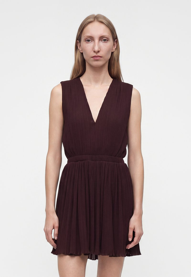 Femme aux cheveux blonds raides portant une robe plissée sans manches violet foncé avec un profond décolleté en V, debout devant un fond blanc uni.