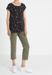 Černý květinový top s krátkým rukávem, zdobený červenými a zelenými vzory, spárovaný se světle olivovými zkrácenými kalhotami a bílými teniskami na jednobarevném pozadí.