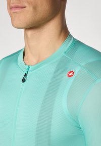 Türkisfarbene Radtrikot mit einem Viertel-Reißverschluss und strukturiertem Stoff. Verfügt über ein kleines rotes Logo auf der linken Schulter. Aerodynamische Passform.