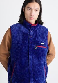 Blue fleece vest with a textured surface, high collar, red accents, and a zippered pocket. Layered over a brown long-sleeve shirt.