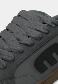 Γκρι αθλητικά παπούτσια με υφαντό ύφασμα, επίπεδα γκρι κορδόνια και μαύρη λογότυπο στη πλευρά, με σόλα από καουτσούκ σε καφέ απόχρωση.