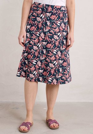 Vrouw die een marineblauwe rok draagt met roze en witte blaadjesprint en paarse slippers, staand op een lichtgrijze vloer.