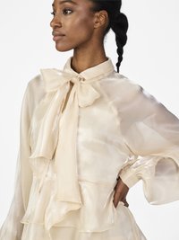 Vrouw in een doorschijnende ivoorkleurige blouse met een grote strik bij de kraag, poseert met haar hand op de heup, kijkt opzij.