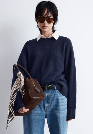 Navy knitted sweater with ribbed cuffs, ivory collared shirt underneath, paired with light blue jeans and a brown suede bag adorned with a patterned scarf.