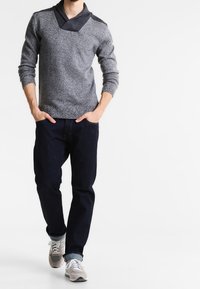 Grauer, strukturierter Pullover mit einem ausgefallenen breiten Kragen, kombiniert mit dunkelblauen Jeans und grauen Sneakers. Lässiges Outfit mit modernen Akzenten.