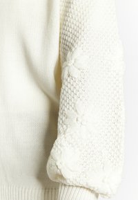Cremefarbener Strickpullover mit strukturiertem Körper, geripptem Saum und bestickten Blumenmotiven an den Ärmeln. Weicher Stoff, lockere Passform.