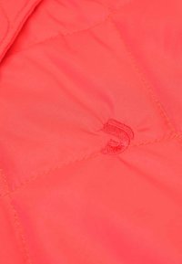 Tissu matelassé de couleur corail vif avec des coutures visibles, présentant un petit logo brodé dans une teinte assortie. Texture lisse.