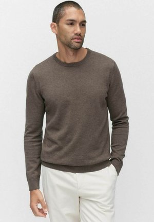 Pull marron avec un col rond, des manches longues et des bords côtelés. Fabriqué à partir d'un matériau doux, présenté avec un pantalon de couleur claire.