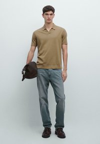 Koszulka polo z krótkim rękawem w jasnobrązowym kolorze, zestawiona z wyblakłymi niebieskimi dżinsami. Model trzyma ciemnobrązową torbę. Buty są brązowe, skórzane, z sznurówkami.