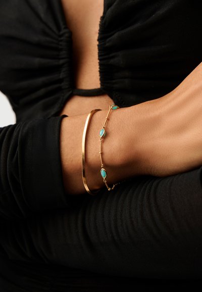 Δύο χρυσά βραχιόλια σε σταυρωμένα χέρια που φορούν μαύρο ρούχο με ρυτίδες, ένα βραχιόλι απλό και το άλλο με μικρές πέτρες τιρκουάζ.