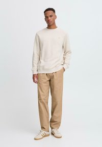 Beżowy sweter z okrągłym dekoltem z ściągaczami, zestawiony z luźnymi, tanowymi spodniami. Model nosi białe sneakersy z beżowymi paskami.
