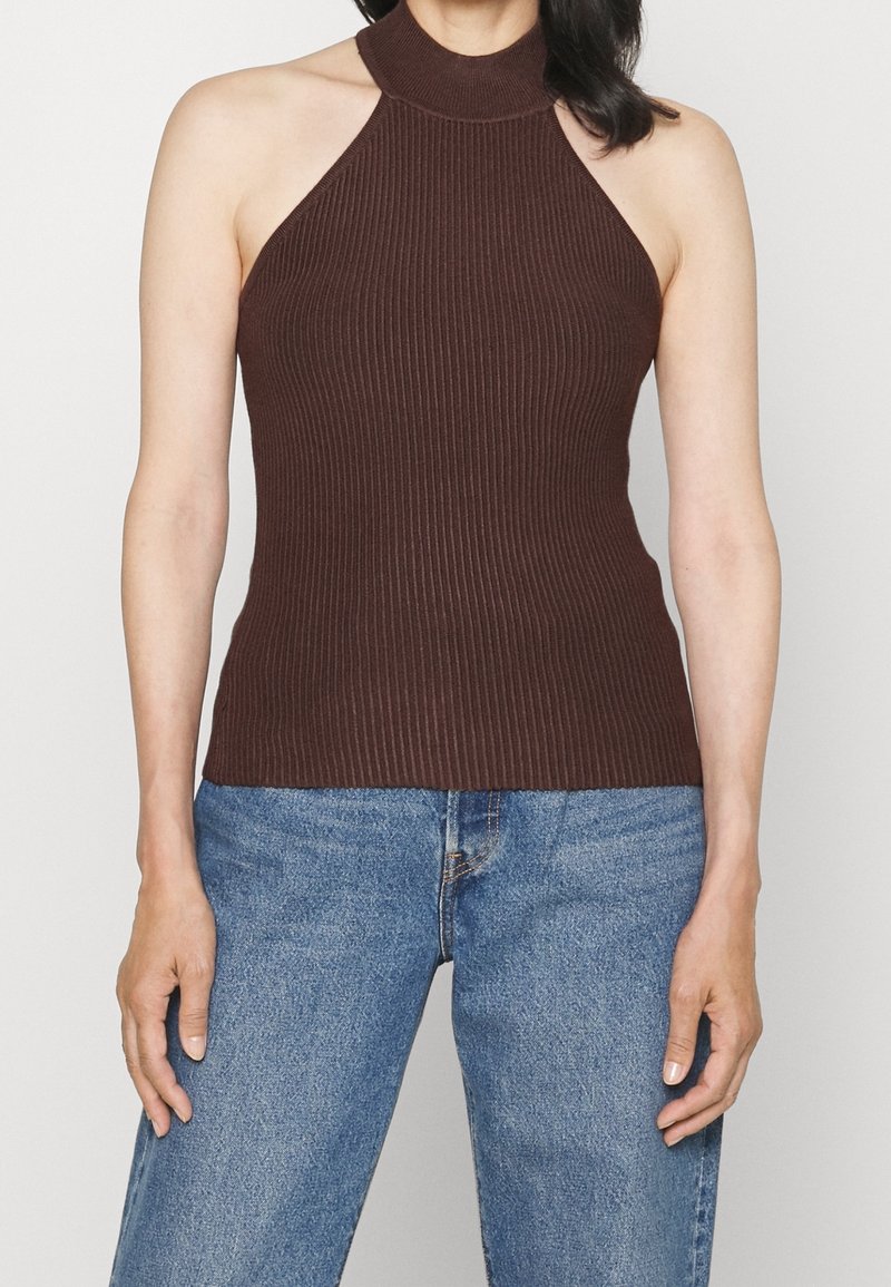 Femme portant un haut sans manches en maille côtelée marron foncé à col licou, associé à un jean en denim bleu, debout devant un fond uni.