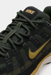 Tenis Nike cu un superior din plasă verde închis, accente negre, logo auriu, suprafețe texturate și un sistem de șireturi distinctiv.