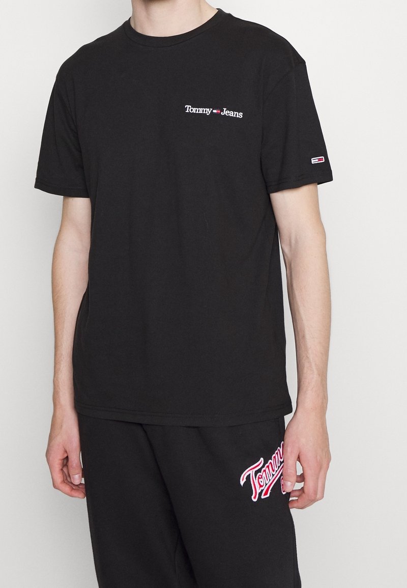 T-shirt en coton noir à manches courtes, arborant un petit logo "Tommy Jeans" rouge et blanc sur la poitrine et un patch de logo sur la manche.