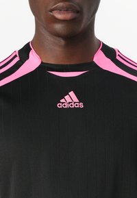 Juodas sportinis marškinėliai su vertikaliomis juostomis, akcentuotas ryškiai rožinėmis petnešomis ir Adidas logotipu. Savybės apvalus apykaklė.