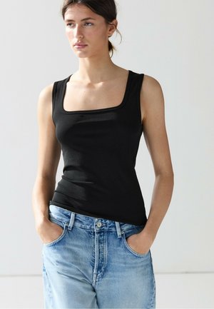 Jeune femme aux cheveux foncés portant un haut noir sans manches à encolure carrée et un jean bleu clair, les mains dans les poches.