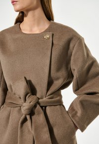 Ruskea fleece-takki, joka on sidottu vyölenkin ympärille, jossa on pyöreä kaula-aukko ja kultainen rintaneula olkapäällä. Pehmeä rakenne ja rento malli.