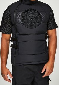 Czarna pikowana kamizelka z wysokim kołnierzem, z teksturowanym czarnym panelem na piersi i dużym okrągłym logo, wyposażona w regulowane paski oraz przednią kieszeń.