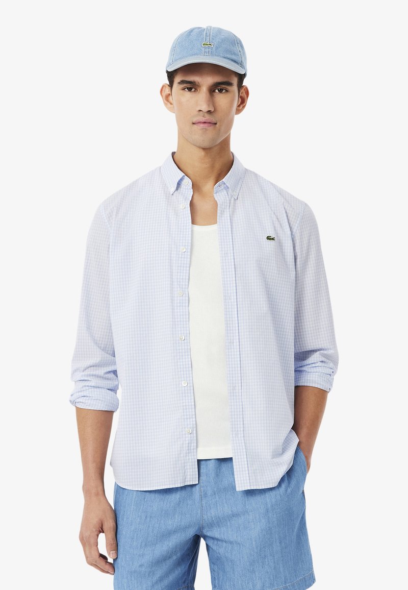 Jeune homme portant une chemise à carreaux bleu clair boutonnée, un débardeur blanc, un short en denim et une casquette de baseball bleu clair.