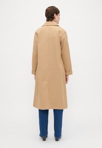 Ilgas kupranugario paltas, pagamintas iš lygios medžiagos, su iškirptėmis ir ilgomis rankovėmis. Dėvimas virš mėlynų džinsų ir tamsių batų.