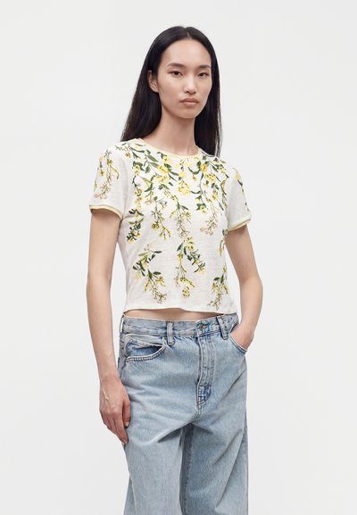 Νεαρή γυναίκα που φοράει λευκό crop top με κοντά μανίκια και κίτρινη κεντητή ανθοδέσμη, και ανοιχτό μπλε τζιν με ψηλή μέση, με τα χέρια στις τσέπες.