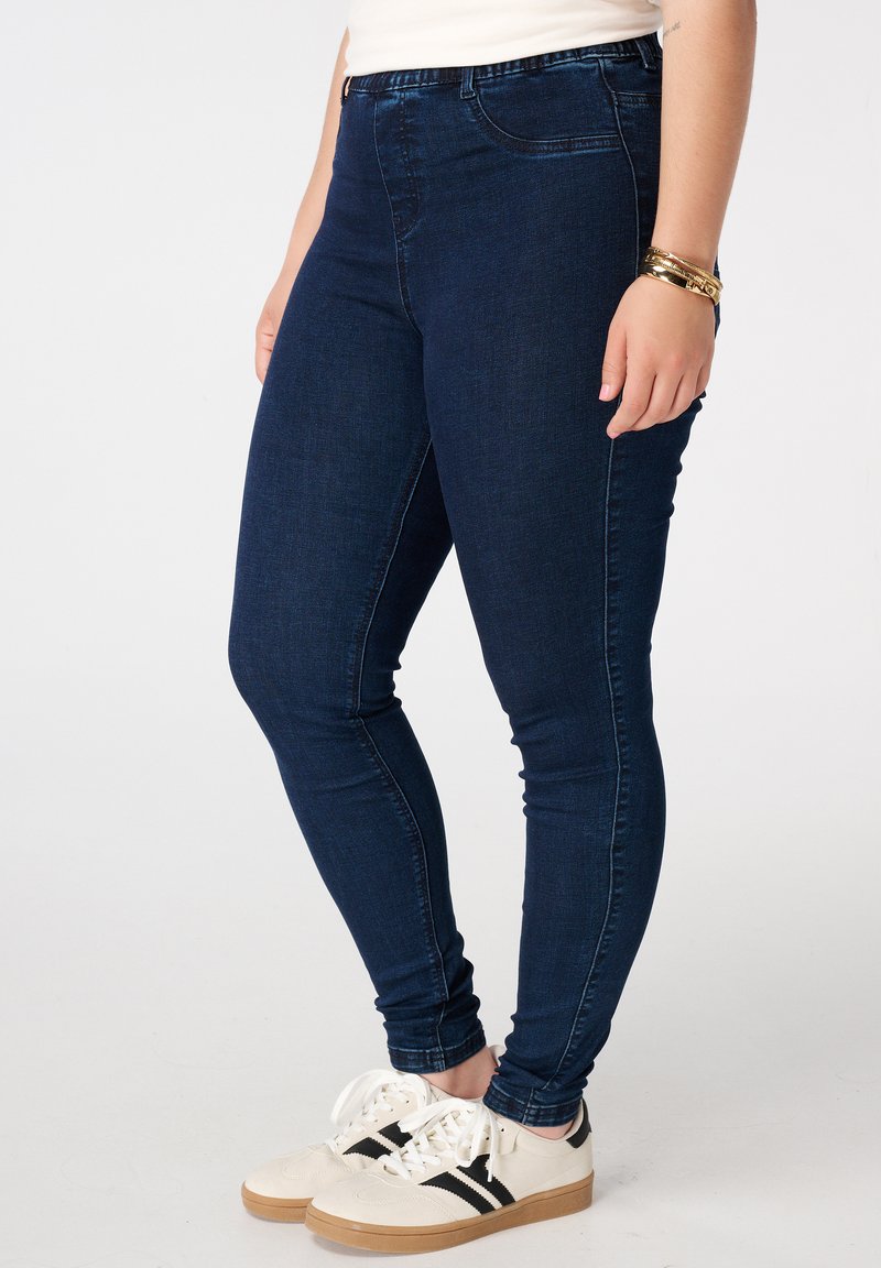 Diepblauwe skinny jeans met een soepele denimtextuur, voorzien van een hoge tailleband en een minimalistisch design, gecombineerd met witte sneakers met zwarte strepen.