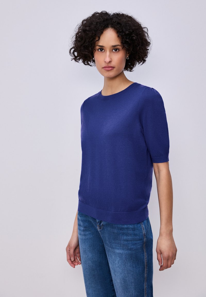 Femme aux cheveux bouclés portant un pull bleu ajusté à manches courtes et un jean bleu, debout devant un fond clair uni.