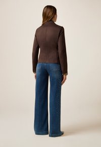 Veste ajustée marron avec un col haut et des manches longues, associée à un jean en denim bleu à jambes larges avec des embellissements sur les côtés.