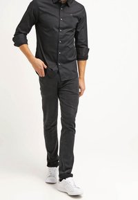 Camisa negra de botones con mangas dobladas, combinada con pantalones ajustados de color gris oscuro y zapatillas blancas. Tela suave y corte entallado.