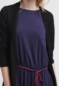 Czarny kardigan nałożony na fioletową sukienkę z przednią kieszenią i bordowym sznurkiem w talii. Gładka tekstura i casualowy design.