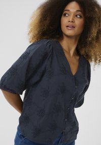 Blusa blu navy con scollo a V, maniche a sbuffo e ricamo floreale testurizzato. Presenta bottoni sul davanti e pizzo sulle maniche.